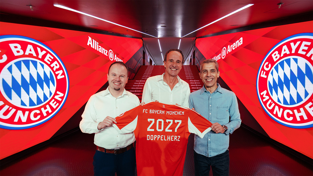 На изображението трима мъже позират с футболна тениска на FC Bayern München, на която е написано „2027 ДОППЕЛХЕРЦ“. На фона се виждат червени стени с логото на клуба, създаващи спортна атмосфера.