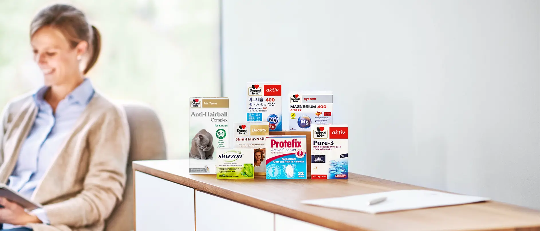 Маса с опаковки на различните брандове на Куайсер Фарма - Допелехерц, Протефикс, Стозон и на заден план жена, която чете